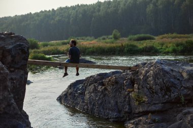 Bir adam nehir kıyısındaki kayaların arasında bir köprüde oturuyor.