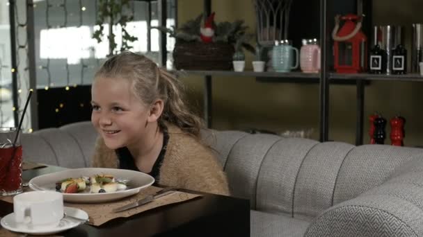 A little girl with blonde hair laughs merrily sitting in a restaurant — Stock Video