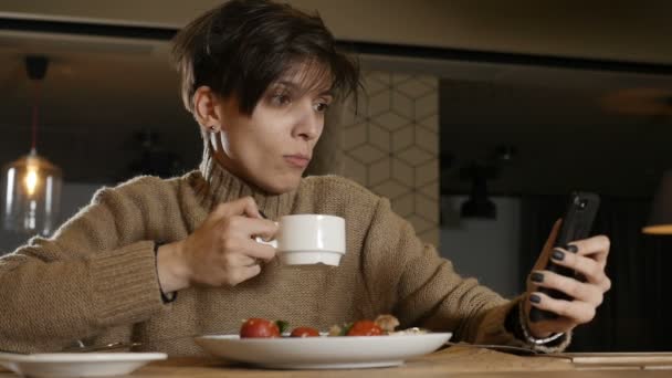 Uma mulher com um corte de cabelo curto olha para o telefone e bebe café — Vídeo de Stock
