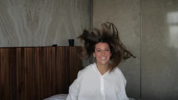 Mooi meisje in een shirt en blote benen vrolijk springt op het bed — Stockvideo