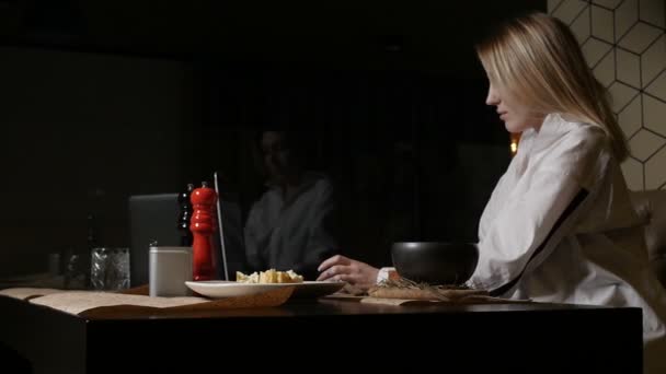 Ξανθιά κοπέλα με λευκό πουκάμισο τρώει ένα πιάτο που κάθεται σε ένα εστιατόριο — Αρχείο Βίντεο