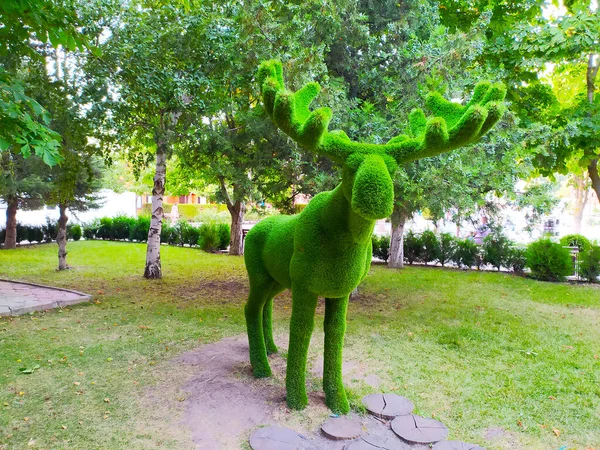 Escultura de un ciervo con cuernos en un parque verde sobre un fondo de árboles. —  Fotos de Stock