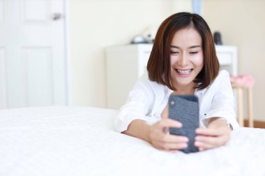 Güzel Asyalı kadın, beyaz tişörtlü, video çağrısı yapan kısa saçlı ya da beyaz yataktaki cep telefonundan kendi portresini çeken mutlu bir yüzü olan.