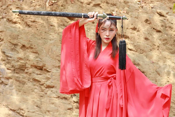 portrait of young and beautiful woman wearing red Chinese warrior costume with black sword, she post holding sword among nature mountain outdoor
