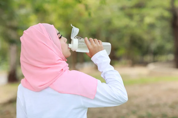 Rear view fitness athlete asian muslim woman drinking water after work out exercising. Young cute girl standing in sportswear with headphone taking rest after workout outdoor.  Healthy, sport concept