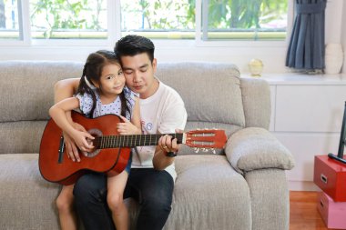 Mutlu Asyalı baba kızına oturma odasındaki gri koltukta gülen yüzlü akustik gitar çalmayı öğretiyor.)