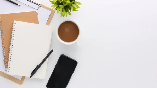 Üzerinde bilgisayar ve ofis malzemeleri olan beyaz manzaralı ahşap ofis masası. Boş defter, klavye, yeşil yaprak ve kahve fincanı olan düz bir çalışma masası. Reklam içeriğiniz için alanı kopyalayın.