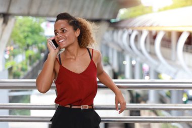 Şehir merkezinde dikilen günlük elbiseli başarılı beyaz iş kadını, gülen yüzlü cep telefonu kullanıyor şehir ve gökyüzü geçmişi ile