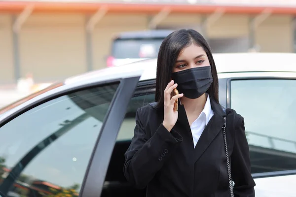 Young asian healthy woman in business black suit with protect mask for healthcare get off automobile and using smartphone. New normal and social distancing concept