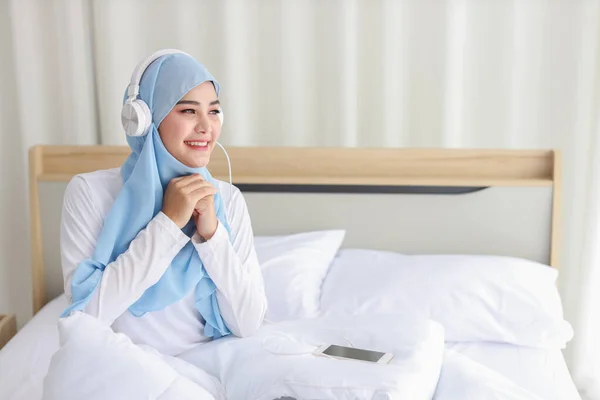 美丽的亚洲穆斯林女性穿着睡衣在手机上观看在线故事的画像 躺在床上 与无线网络相连 年轻可爱的戴头巾的女孩听智能手机里的音乐 — 图库照片