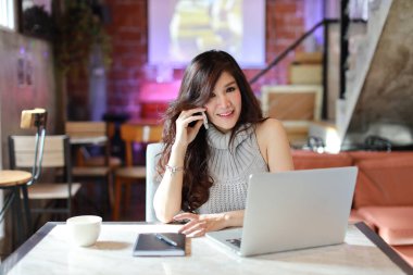 Güzel ve çekici Asyalı kadın, kafeteryada cep telefonu ve dizüstü bilgisayar kullanırken evrak işleriyle uğraşan gri günlük elbiseli uzun saçlı.