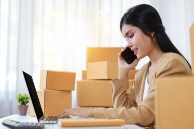 Yan görünüm genç Asyalı akıllı ve mutlu kadın evde dizüstü bilgisayar ve akıllı telefonla çalışan online alışveriş siparişi ve kutu ambalajı olan (yeni normal konsept))