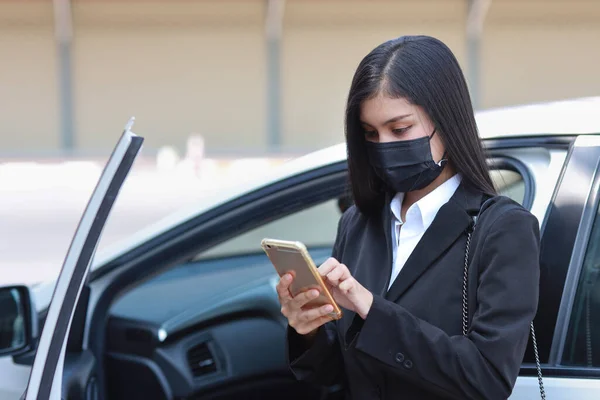 Young asian healthy woman in business black suit with protect mask for healthcare get off automobile and using smartphone. New normal and social distancing concept