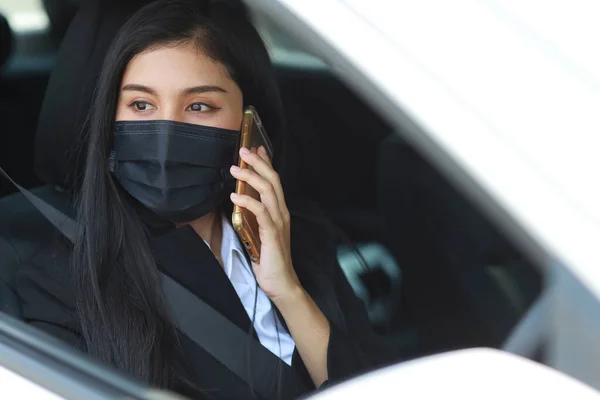 Young asian healthy woman in business black suit with protect mask for healthcare in automobile and using smartphone and driving car. New normal and social distancing concept