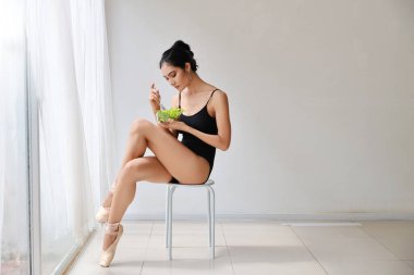 Güzel, sağlıklı ve sportif Asyalı genç bir kadın salata kasesi tutuyor ve bale çalışmasından sonra yemek yiyor ve mutlu bir gülümsemeyle oturuyor.