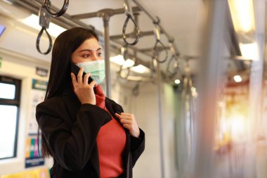 Yeni normal covid-19 salgını. Trendeki insanlardan virüs kapmasını önlemek için maske takan Asyalı genç bir kadın. Trende akıllı telefon iletişimi kullanıyor.