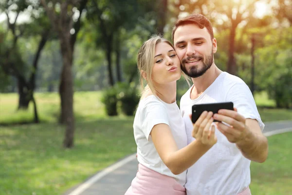 年轻的高加索夫妇男女数码成瘾的社会时代 使用手机视频通话 并向公园里有绿树背景的人发送亲吻 技术和生活方式概念 — 图库照片