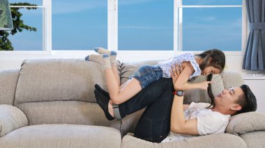 Beyaz tişörtlü mutlu Asyalı bekar baba ve mavi kot, Asyalı kızını oturma odasında gri koltukta gülümseyen yüzüyle yatarken fırlatıyor.)