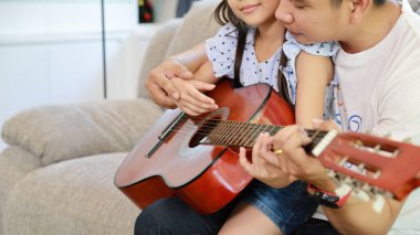 Mutlu Asyalı baba, kızına oturma odasındaki gri koltukta akustik gitar çalmayı öğretiyor. Gülen ve gülümseyen yüzüyle.)