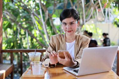 Sağlık hizmetleri için yüz maskesi takan Asyalı genç bir kadın kafeteryada oturuyor, akıllı telefon kullanıyor ve dizüstü bilgisayar üzerinde çalışıyor. Yeni normal ve sosyal uzaklık kavramı