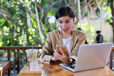 Sağlık hizmetleri için yüz maskesi takan Asyalı genç bir kadın kafeteryada oturuyor, akıllı telefon kullanıyor ve dizüstü bilgisayar üzerinde çalışıyor. Yeni normal ve sosyal uzaklık kavramı