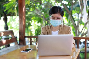Genç Asyalı kadın günlük kıyafetlerini giyip sağlık için maske takıyor, kafeteryada oturuyor ve akıllı telefon kullanıyor ve dizüstü bilgisayar üzerinde çalışıyor. Yeni normal ve sosyal uzaklık kavramı