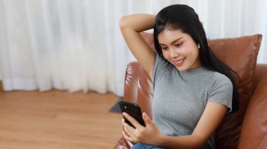 Genç, zeki ve aktif Asyalı kadın oturma odasında koltukta oturuyor. Kablosuz kulaklıklı akıllı telefon kullanıyor ve gülümseyen bir yüz takınıyor.