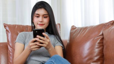 Genç, zeki ve aktif Asyalı kadın oturma odasında koltukta oturuyor. Kablosuz kulaklıklı akıllı telefon kullanıyor ve gülümseyen bir yüz takınıyor.