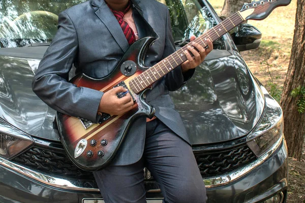 Yol kenarındaki arabanın kaputunda oturan genç bir adam elektro gitar çalıyor. Müzik aletleri, tutku ve hobi kavramları..