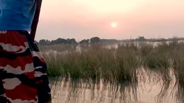 Video görüntüleri, nehir kıyısındaki kayıkçının arka planında yüzen bir tekneden çekilen çarpıcı bir günbatımını gösteriyor. Batan güneşin ve ağaçların kırmızı renkleri nehir suyuna çok güzel yansıyor..