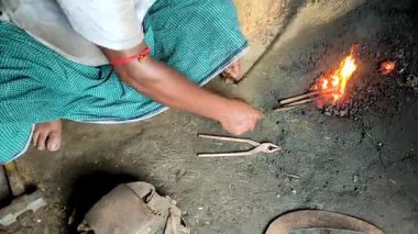 Geleneksel bir demirci, maşa ve basit aletlerle metali ısıtırken küçük bir kömür ocağının yanında çalışıyor. Kırsal işçiliğin ve geleneksel metal dövme tekniklerinin vurgulanması.