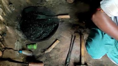 Geleneksel bir demirci, maşa ve basit aletlerle metali ısıtırken küçük bir kömür ocağının yanında çalışıyor. Kırsal işçiliğin ve geleneksel metal dövme tekniklerinin vurgulanması.