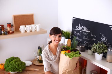 Sebzeli alışveriş poşeti taşıyan genç bir kadın mutfakta duruyor.