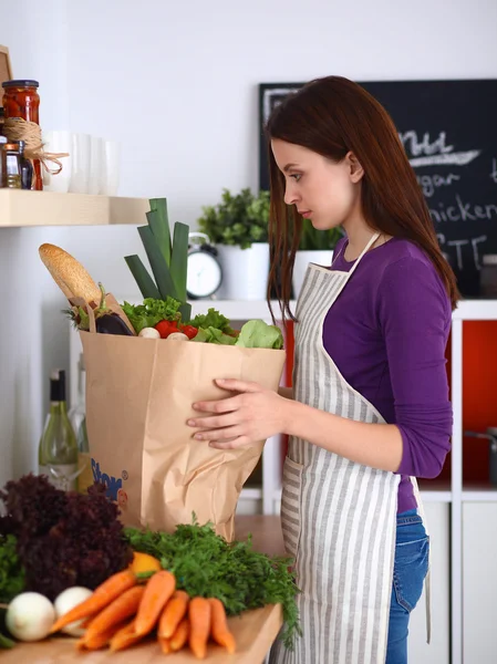 Mujer joven de pie en su cocina cerca del escritorio con bolsas de compras —  Fotos de Stock