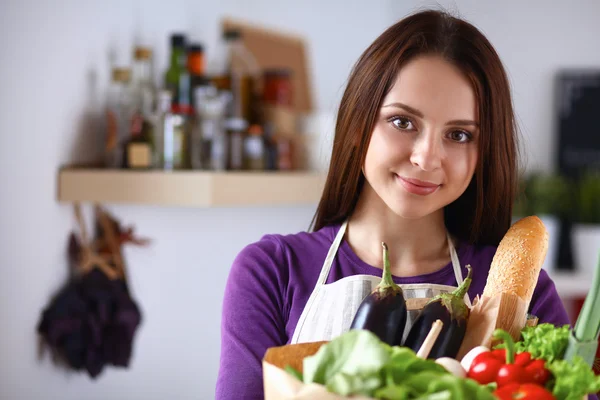 Mujer joven sosteniendo bolsa de la compra de comestibles con verduras de pie en la cocina —  Fotos de Stock
