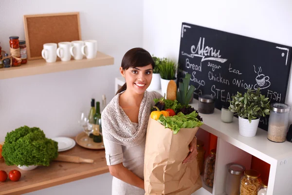 Mujer joven sosteniendo bolsa de la compra de comestibles con verduras de pie en la cocina —  Fotos de Stock