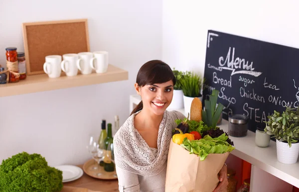 Mujer joven sosteniendo bolsa de la compra de comestibles con verduras de pie en la cocina —  Fotos de Stock