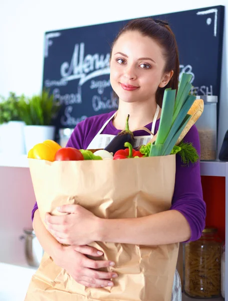 Mujer joven sosteniendo bolsa de la compra de comestibles con verduras de pie en la cocina —  Fotos de Stock