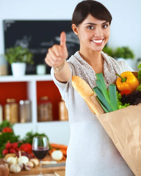 Mujer joven sosteniendo la bolsa de la compra de comestibles con verduras de pie en la cocina y mostrando ok —  Fotos de Stock