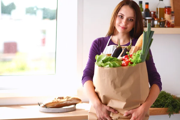 Mujer joven sosteniendo bolsa de la compra de comestibles con verduras de pie en la cocina —  Fotos de Stock