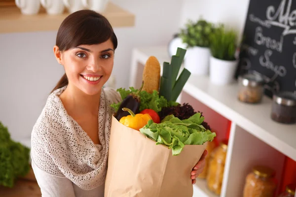 Mujer joven sosteniendo bolsa de la compra de comestibles con verduras de pie en la cocina —  Fotos de Stock