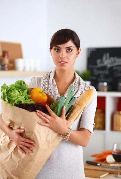 Mujer joven sosteniendo bolsa de la compra de comestibles con verduras de pie en la cocina —  Fotos de Stock