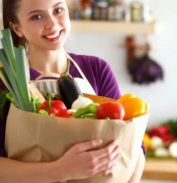 Mujer joven sosteniendo bolsa de la compra de comestibles con verduras de pie en la cocina —  Fotos de Stock
