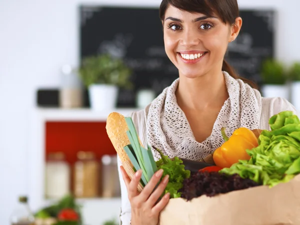 Mujer joven sosteniendo bolsa de la compra de comestibles con verduras de pie en la cocina —  Fotos de Stock