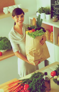 Sebzeli alışveriş poşeti taşıyan genç bir kadın mutfakta duruyor.
