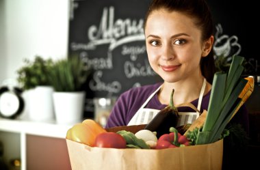 Sebzeli alışveriş poşeti taşıyan genç bir kadın mutfakta duruyor.