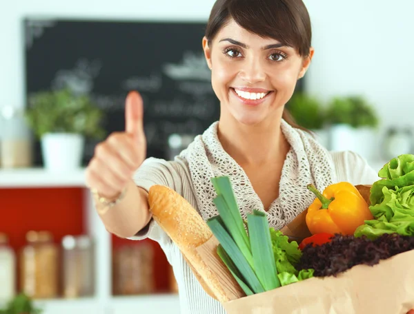 Mujer joven sosteniendo la bolsa de la compra de comestibles con verduras de pie en la cocina y mostrando ok —  Fotos de Stock