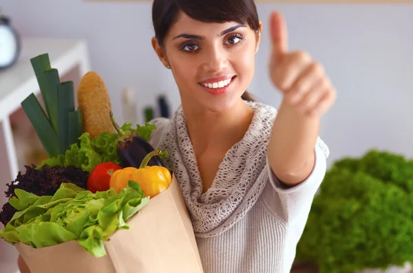 Mujer joven sosteniendo la bolsa de la compra de comestibles con verduras de pie en la cocina y mostrando ok — Foto de Stock