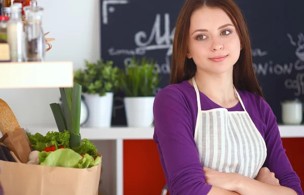 Uma jovem mulher de pé em sua cozinha com saco de mantimentos no fundo . — Fotografia de Stock