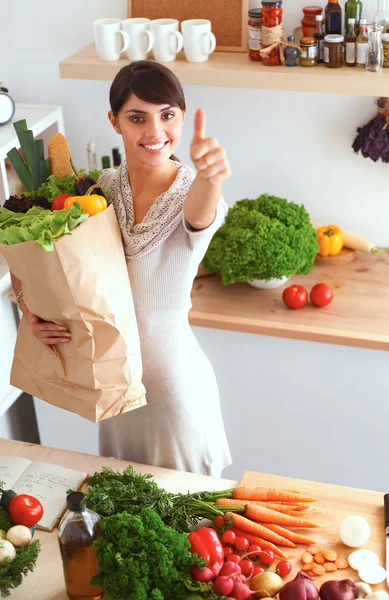 Mujer joven sosteniendo la bolsa de la compra de comestibles con verduras de pie en la cocina y mostrando ok —  Fotos de Stock